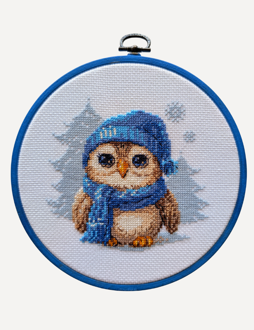 Cuddly Owl cross-stitch kit showing an owl in blue hat and scarf standing in snow, winter embroidery design by Aine.