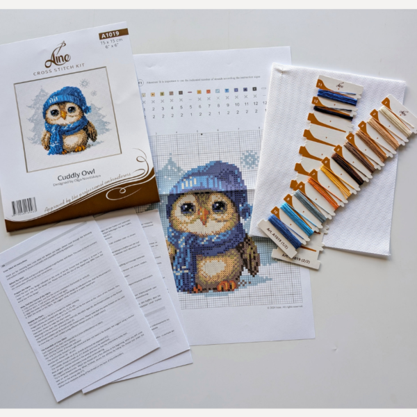 Cuddly Owl cross-stitch kit showing an owl in blue hat and scarf standing in snow, winter embroidery design by Aine.