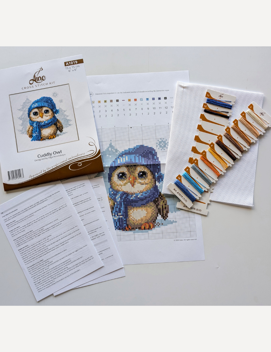 Cuddly Owl cross-stitch kit showing an owl in blue hat and scarf standing in snow, winter embroidery design by Aine.