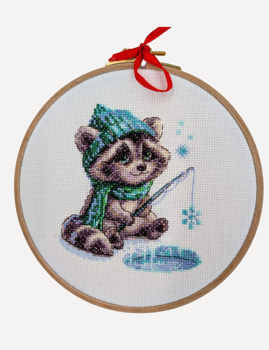 Aine A1017 Curious Raccoon Cross Stitch Kit – counted cross stitch design featuring a raccoon in a green scarf and hat, sitting beside a frozen pond, fishing for snowflakes; includes 14 ct white Zweigart Aida fabric, 15 Anchor stranded cotton threads, needle, color chart, and instructions; finished size 15 x 15 cm (6 x 6 inches).