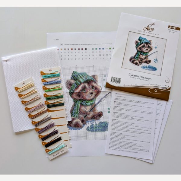 Aine A1017 Curious Raccoon Cross Stitch Kit – counted cross stitch design featuring a raccoon in a green scarf and hat, sitting beside a frozen pond, fishing for snowflakes; includes 14 ct white Zweigart Aida fabric, 15 Anchor stranded cotton threads, needle, color chart, and instructions; finished size 15 x 15 cm (6 x 6 inches).