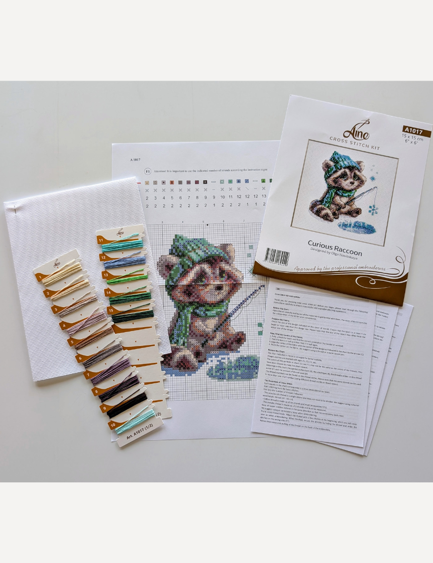 Aine A1017 Curious Raccoon Cross Stitch Kit – counted cross stitch design featuring a raccoon in a green scarf and hat, sitting beside a frozen pond, fishing for snowflakes; includes 14 ct white Zweigart Aida fabric, 15 Anchor stranded cotton threads, needle, color chart, and instructions; finished size 15 x 15 cm (6 x 6 inches).