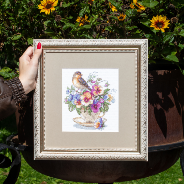A completed cross-stitch of a small brown bird perched on a teacup filled with colorful pansies in shades of purple, blue, orange, and yellow, stitched on white Aida fabric.