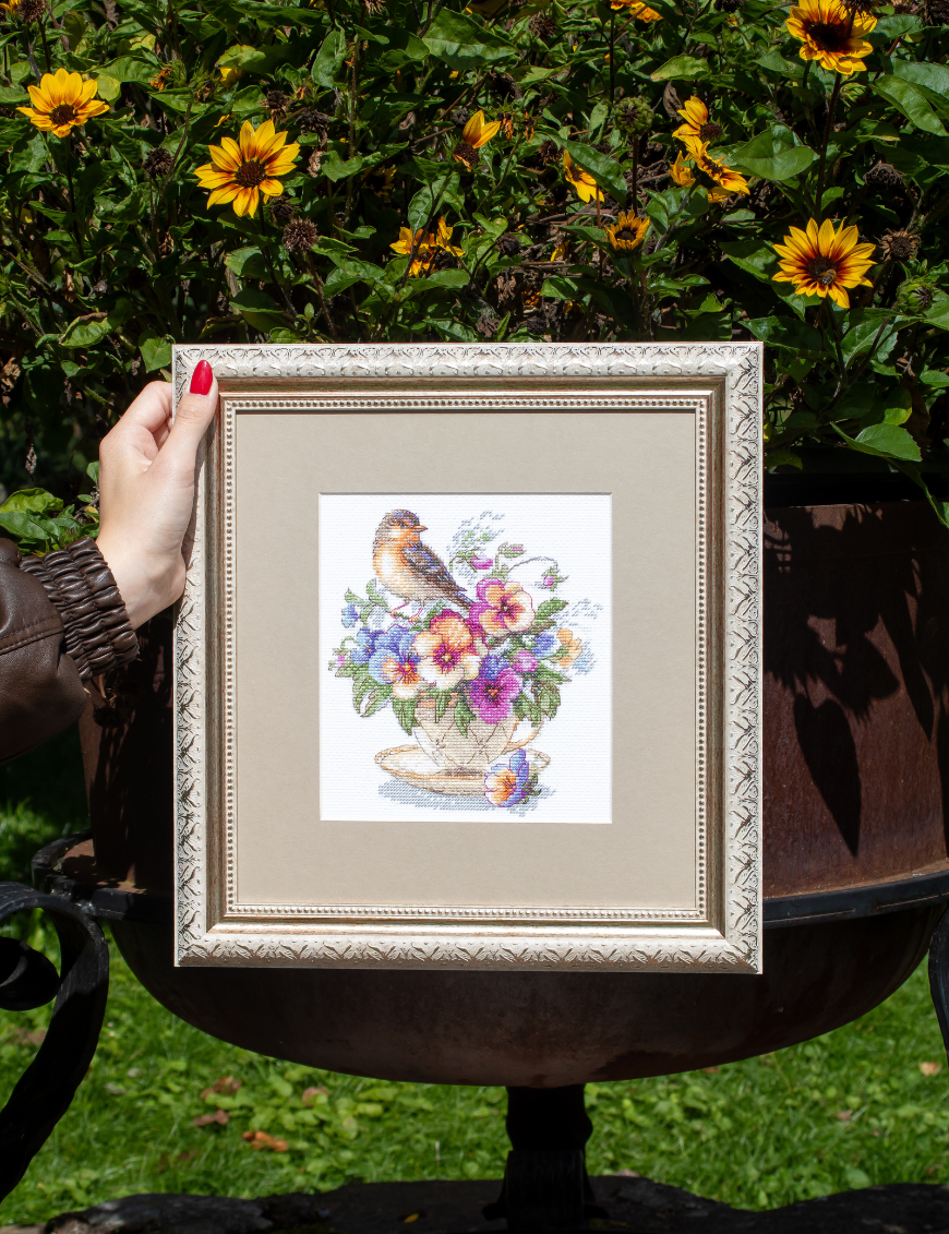 A completed cross-stitch of a small brown bird perched on a teacup filled with colorful pansies in shades of purple, blue, orange, and yellow, stitched on white Aida fabric.