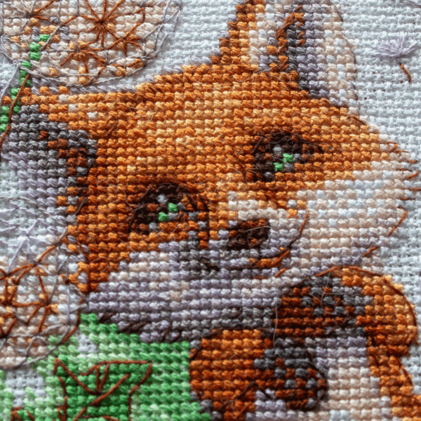 A completed cross-stitch of a smiling red fox lying among dandelion puffs, with seeds floating away, stitched in bright orange, green, and soft lavender tones on white Aida fabric.