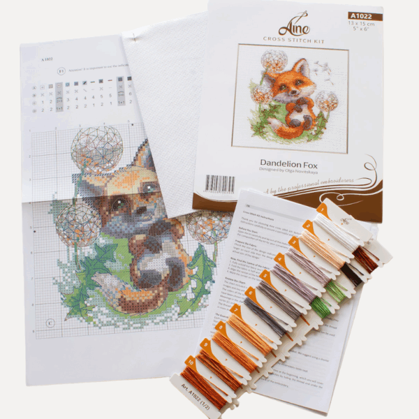 A completed cross-stitch of a smiling red fox lying among dandelion puffs, with seeds floating away, stitched in bright orange, green, and soft lavender tones on white Aida fabric.