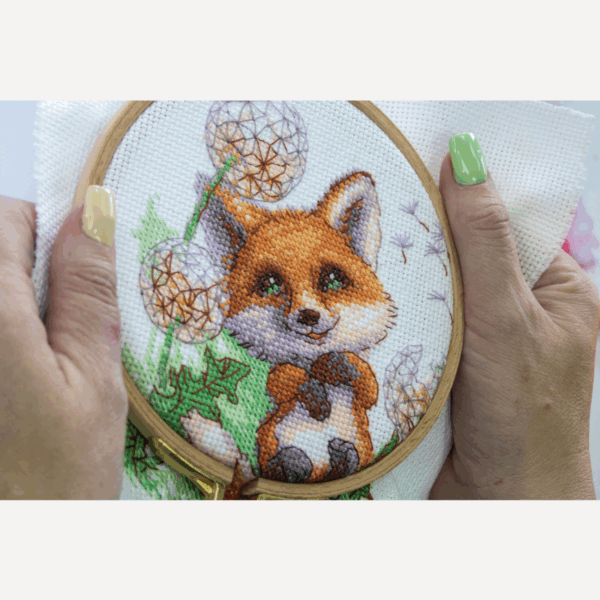 A completed cross-stitch of a smiling red fox lying among dandelion puffs, with seeds floating away, stitched in bright orange, green, and soft lavender tones on white Aida fabric.