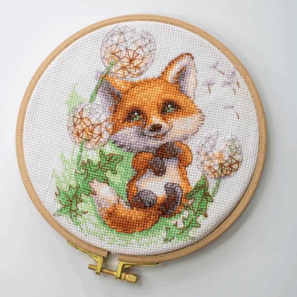 A completed cross-stitch of a smiling red fox lying among dandelion puffs, with seeds floating away, stitched in bright orange, green, and soft lavender tones on white Aida fabric.