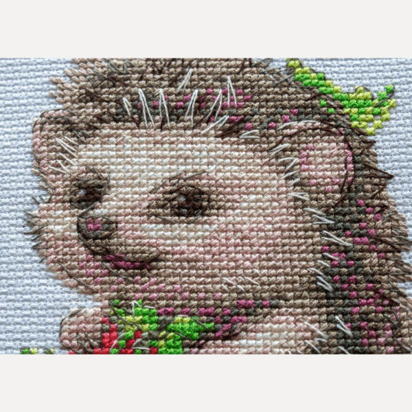 A completed cross-stitch of a smiling hedgehog holding two bright red strawberries, with a small ladybug above, stitched in warm browns, pinks, and reds on white Aida fabric.