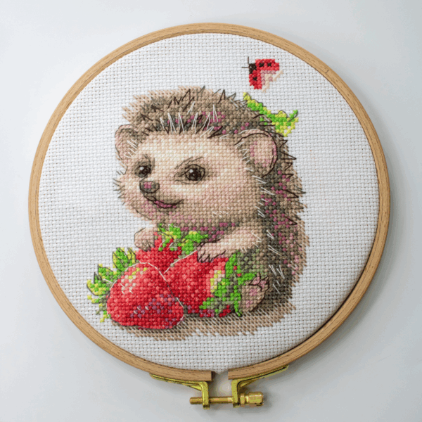 A completed cross-stitch of a smiling hedgehog holding two bright red strawberries, with a small ladybug above, stitched in warm browns, pinks, and reds on white Aida fabric.