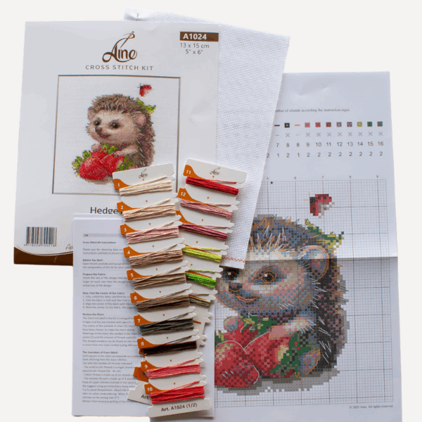 A completed cross-stitch of a smiling hedgehog holding two bright red strawberries, with a small ladybug above, stitched in warm browns, pinks, and reds on white Aida fabric.