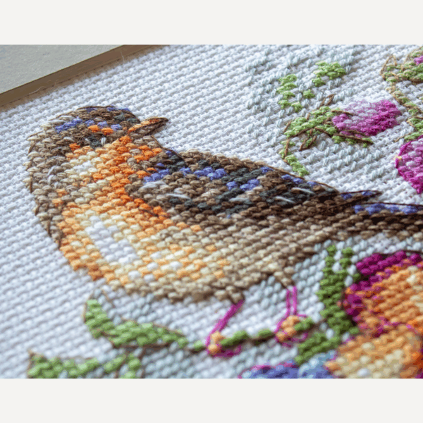 A completed cross-stitch of a small brown bird perched on a teacup filled with colorful pansies in shades of purple, blue, orange, and yellow, stitched on white Aida fabric.
