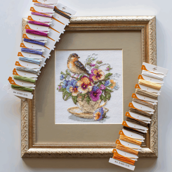 A completed cross-stitch of a small brown bird perched on a teacup filled with colorful pansies in shades of purple, blue, orange, and yellow, stitched on white Aida fabric.