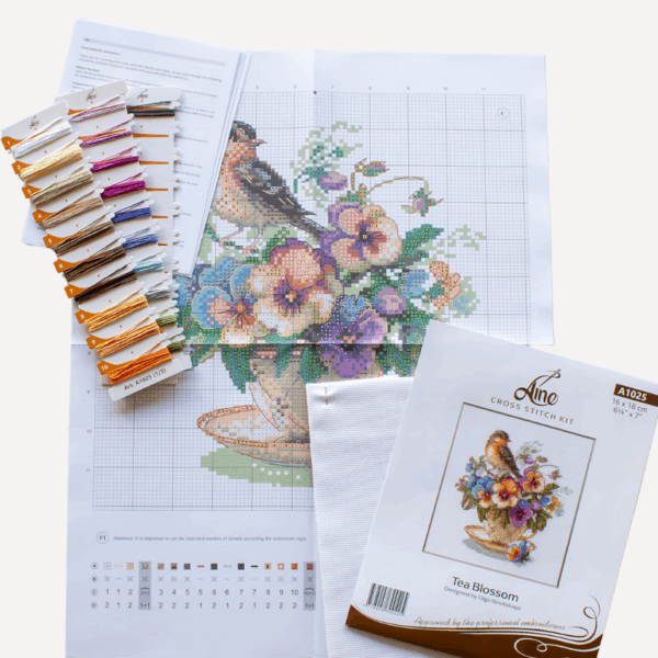 A completed cross-stitch of a small brown bird perched on a teacup filled with colorful pansies in shades of purple, blue, orange, and yellow, stitched on white Aida fabric.