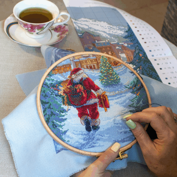 A completed cross-stitch of Santa Claus walking through a snowy village square with a glowing Christmas tree, surrounded by decorated houses and snowy mountains in the background.