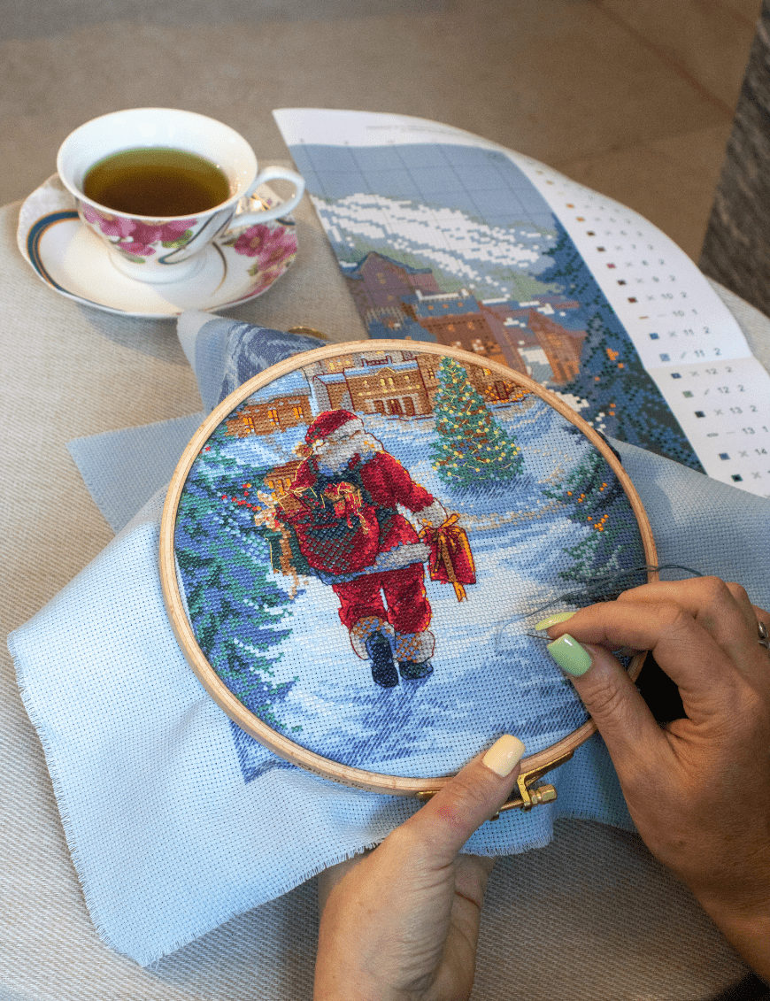 A completed cross-stitch of Santa Claus walking through a snowy village square with a glowing Christmas tree, surrounded by decorated houses and snowy mountains in the background.