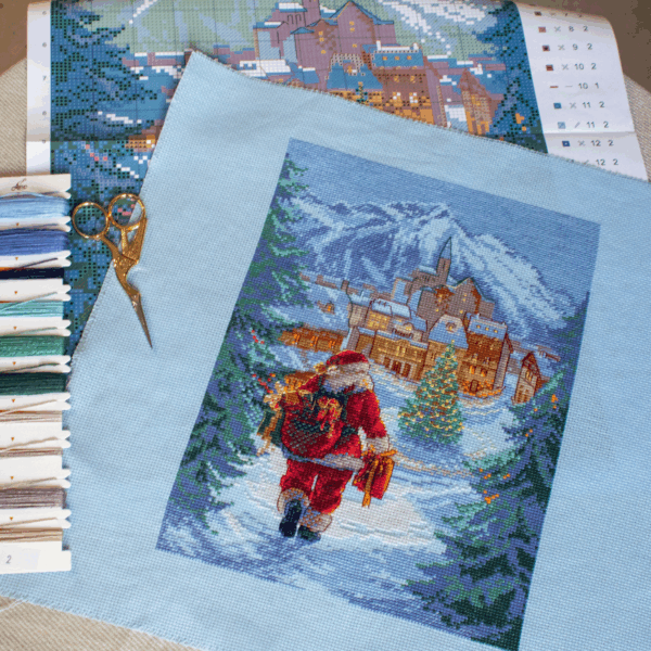 A completed cross-stitch of Santa Claus walking through a snowy village square with a glowing Christmas tree, surrounded by decorated houses and snowy mountains in the background.