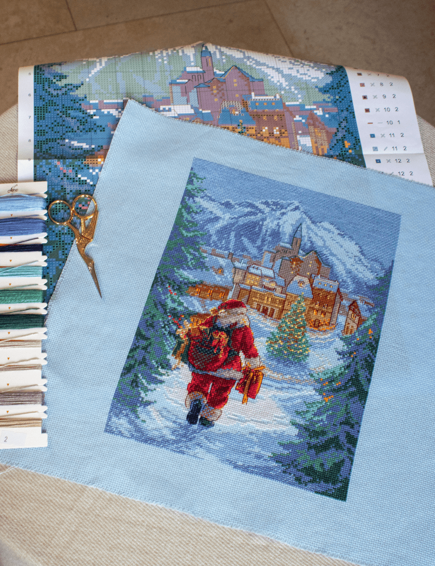 A completed cross-stitch of Santa Claus walking through a snowy village square with a glowing Christmas tree, surrounded by decorated houses and snowy mountains in the background.