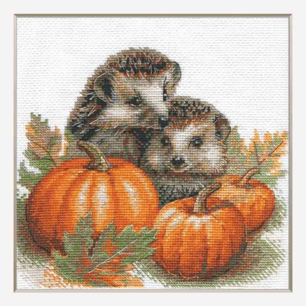 A completed cross-stitch of two hedgehogs nestled among pumpkins and autumn leaves, stitched in warm browns, oranges, and greens on white Aida fabric.