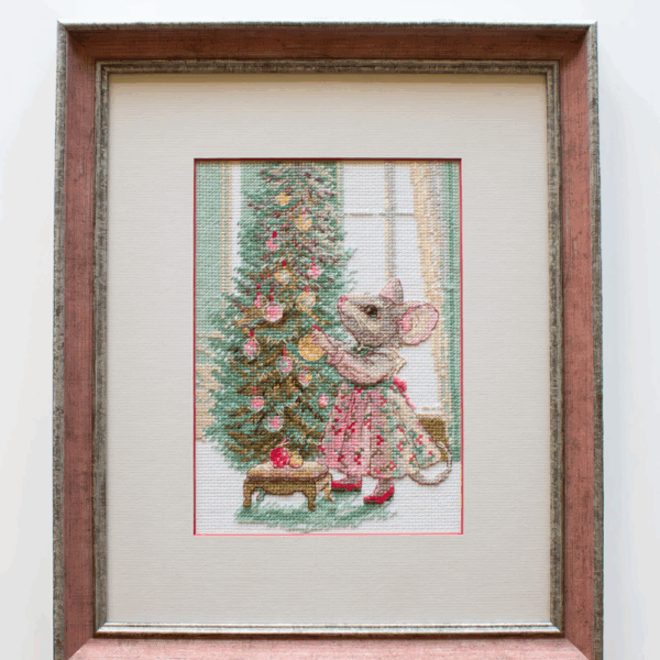 A completed cross-stitch of a mouse in a festive dress decorating a Christmas tree with ornaments, stitched in warm reds, greens, and golds on white Aida fabric.