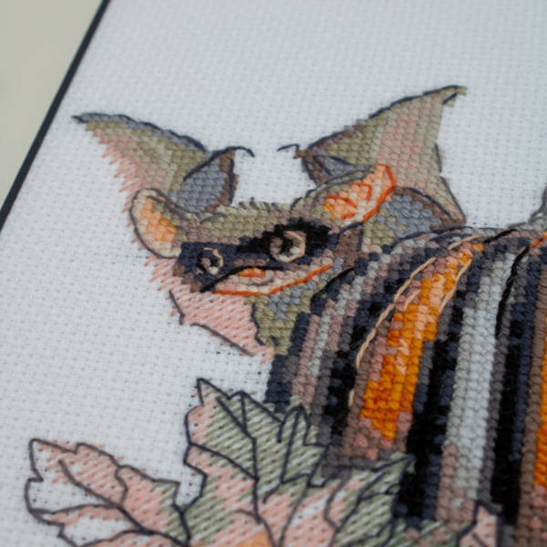 A completed cross-stitch of bats and pumpkins in a Halloween theme, with striped and checkered pumpkins, autumn leaves, and a bat in a witch hat holding a wand.