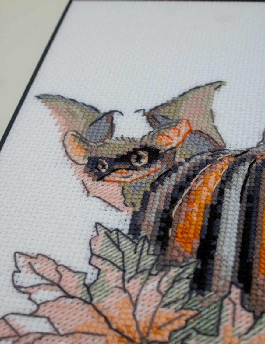 A completed cross-stitch of bats and pumpkins in a Halloween theme, with striped and checkered pumpkins, autumn leaves, and a bat in a witch hat holding a wand.