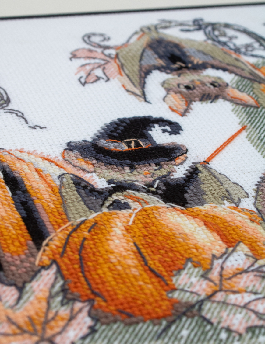 A completed cross-stitch of bats and pumpkins in a Halloween theme, with striped and checkered pumpkins, autumn leaves, and a bat in a witch hat holding a wand.