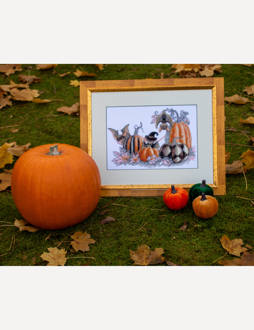 A completed cross-stitch of bats and pumpkins in a Halloween theme, with striped and checkered pumpkins, autumn leaves, and a bat in a witch hat holding a wand.