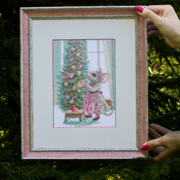 A completed cross-stitch of a mouse in a festive dress decorating a Christmas tree with ornaments, stitched in warm reds, greens, and golds on white Aida fabric.