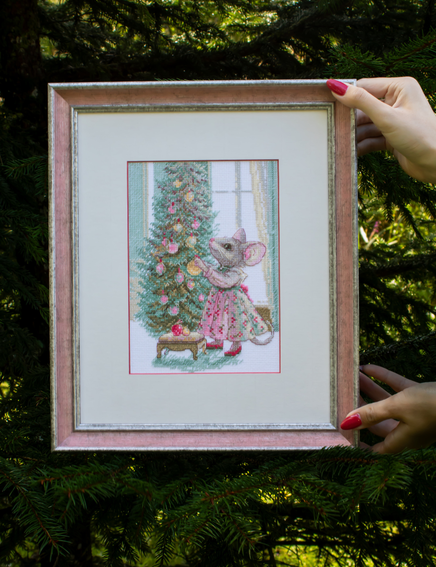 A completed cross-stitch of a mouse in a festive dress decorating a Christmas tree with ornaments, stitched in warm reds, greens, and golds on white Aida fabric.