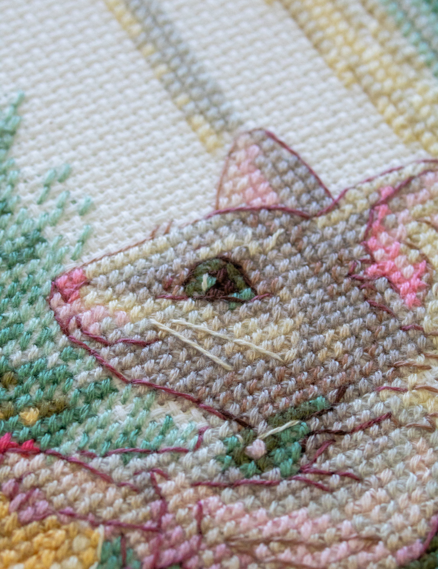 A completed cross-stitch of a mouse in a festive dress decorating a Christmas tree with ornaments, stitched in warm reds, greens, and golds on white Aida fabric.
