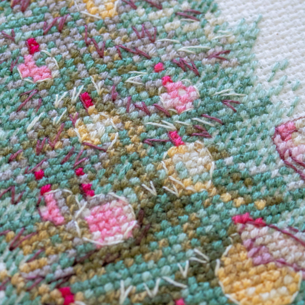 A completed cross-stitch of a mouse in a festive dress decorating a Christmas tree with ornaments, stitched in warm reds, greens, and golds on white Aida fabric.