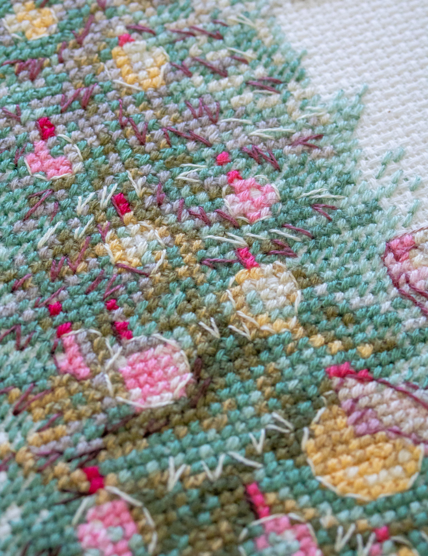 A completed cross-stitch of a mouse in a festive dress decorating a Christmas tree with ornaments, stitched in warm reds, greens, and golds on white Aida fabric.