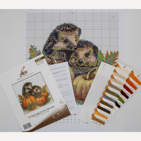 A completed cross-stitch of two hedgehogs nestled among pumpkins and autumn leaves, stitched in warm browns, oranges, and greens on white Aida fabric.
