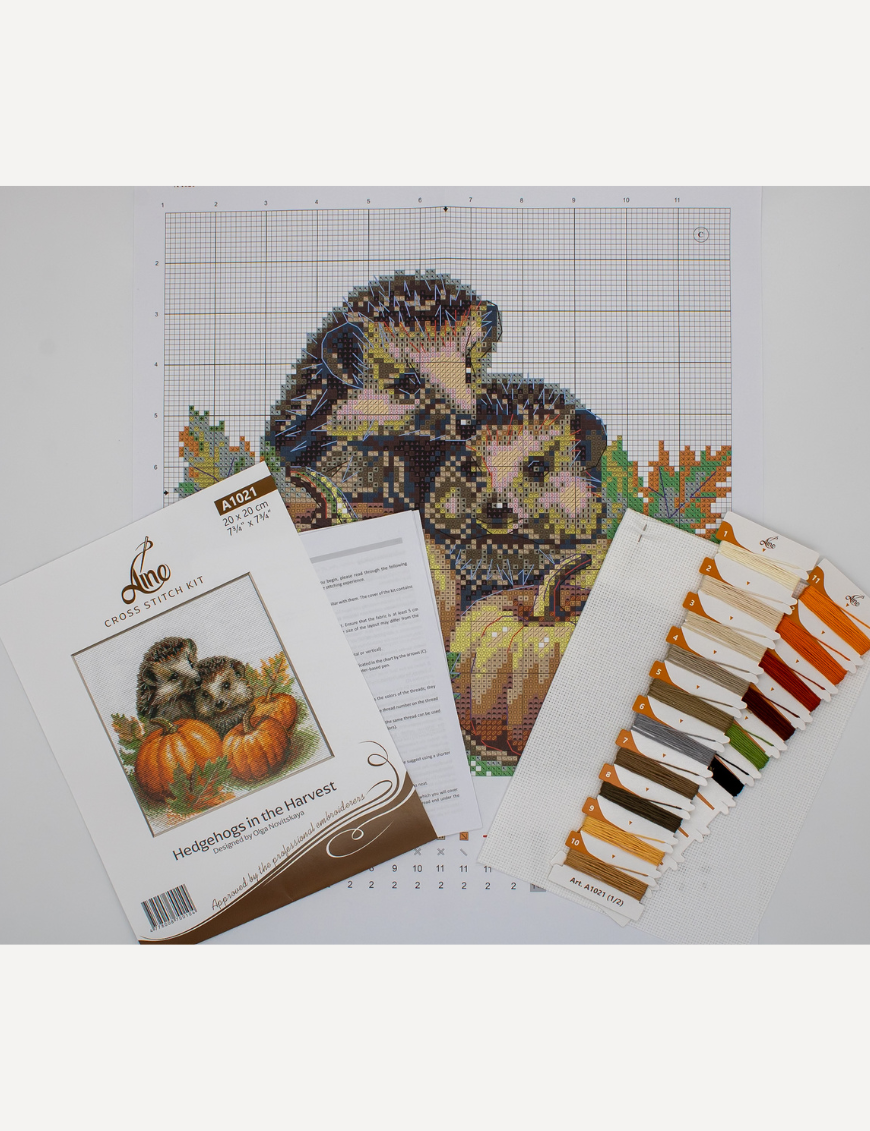 A completed cross-stitch of two hedgehogs nestled among pumpkins and autumn leaves, stitched in warm browns, oranges, and greens on white Aida fabric.