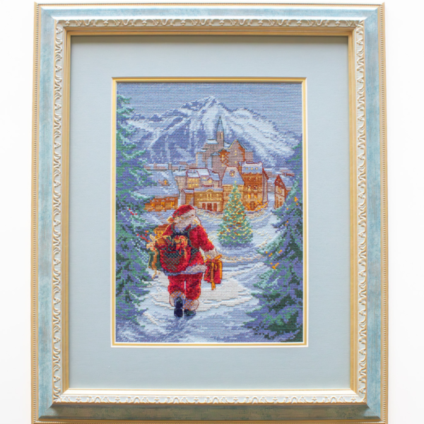 A completed cross-stitch of Santa Claus walking through a snowy village square with a glowing Christmas tree, surrounded by decorated houses and snowy mountains in the background.