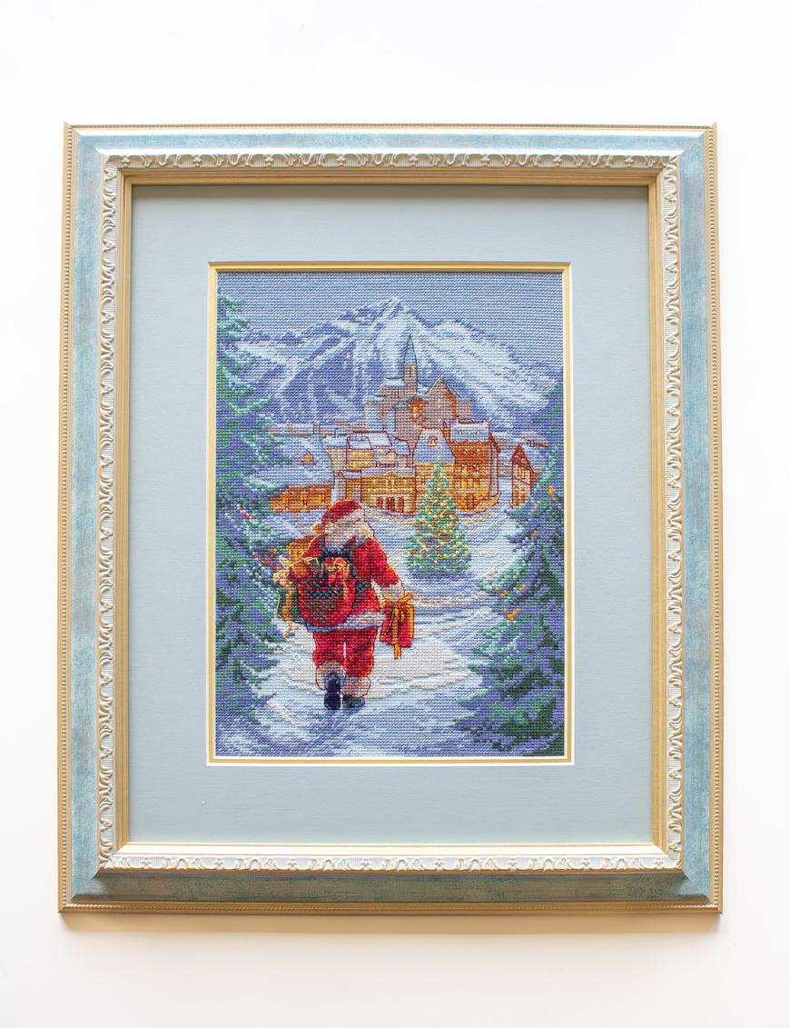 A completed cross-stitch of Santa Claus walking through a snowy village square with a glowing Christmas tree, surrounded by decorated houses and snowy mountains in the background.