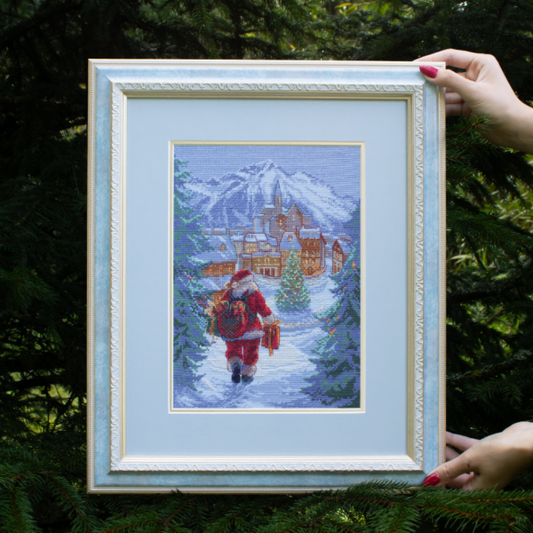 A completed cross-stitch of Santa Claus walking through a snowy village square with a glowing Christmas tree, surrounded by decorated houses and snowy mountains in the background.