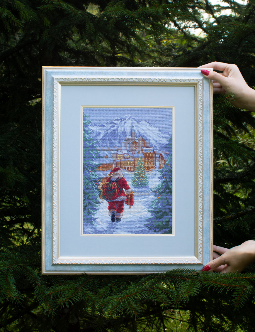 A completed cross-stitch of Santa Claus walking through a snowy village square with a glowing Christmas tree, surrounded by decorated houses and snowy mountains in the background.
