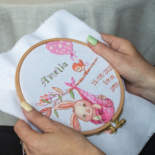 A completed cross-stitch birth sampler featuring a bunny in a pink sling hanging from a branch, with a bird holding a pink heart balloon and space for baby details.