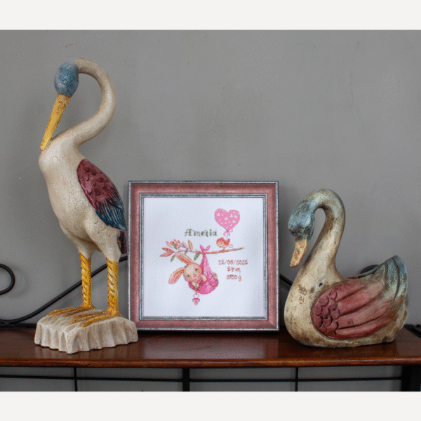 A completed cross-stitch birth sampler featuring a bunny in a pink sling hanging from a branch, with a bird holding a pink heart balloon and space for baby details.