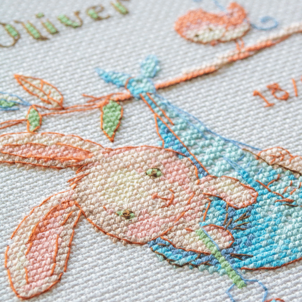 A completed cross-stitch birth sampler featuring a bunny in a blue sling hanging from a branch, with a bird holding a blue heart balloon and space for baby details.