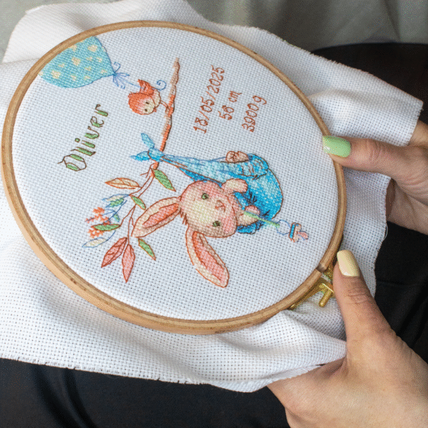A completed cross-stitch birth sampler featuring a bunny in a blue sling hanging from a branch, with a bird holding a blue heart balloon and space for baby details.