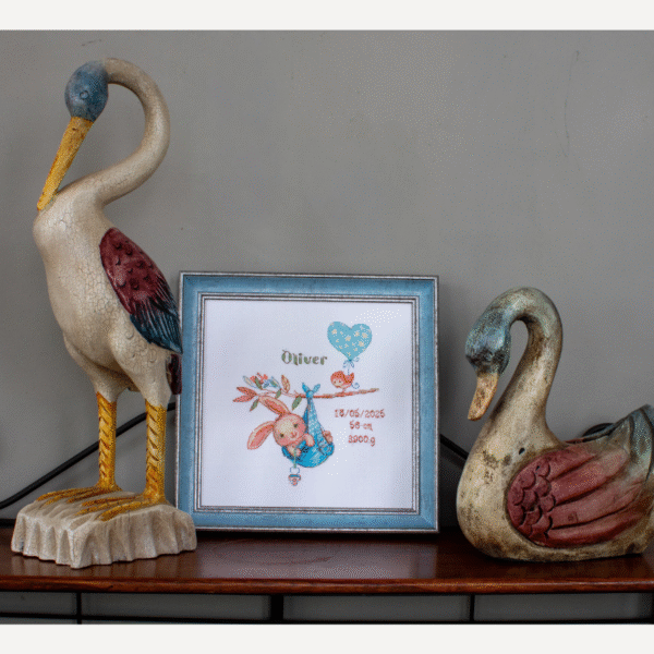 A completed cross-stitch birth sampler featuring a bunny in a blue sling hanging from a branch, with a bird holding a blue heart balloon and space for baby details.