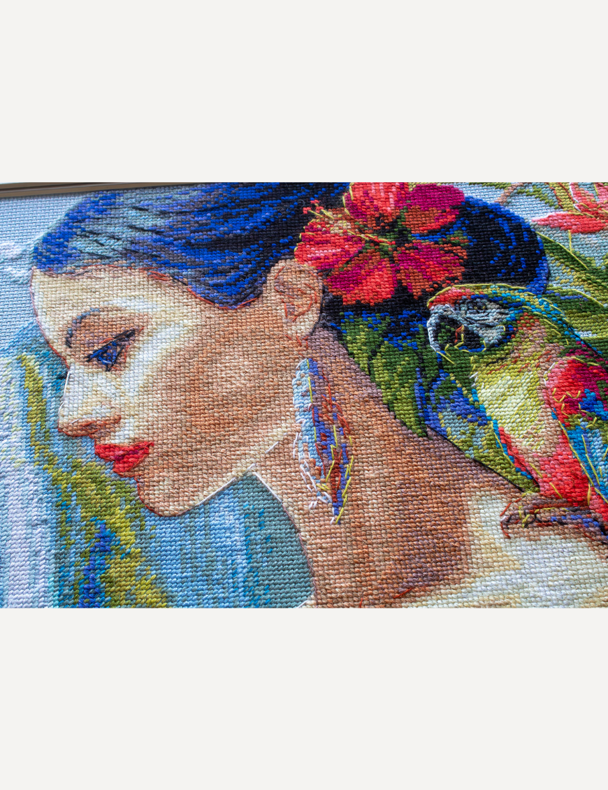 A completed cross-stitch of a woman with red hibiscus flowers in her hair, a colorful parrot on her shoulder, tropical leaves in the foreground, and waterfalls in the background.
