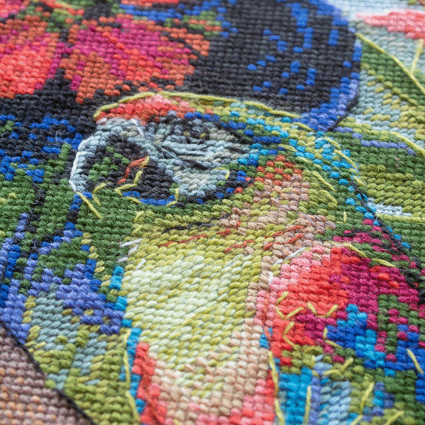 A completed cross-stitch of a woman with red hibiscus flowers in her hair, a colorful parrot on her shoulder, tropical leaves in the foreground, and waterfalls in the background.