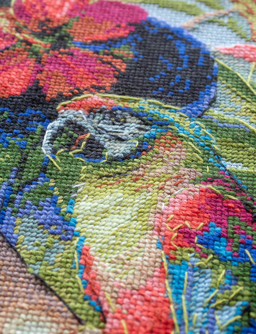 A completed cross-stitch of a woman with red hibiscus flowers in her hair, a colorful parrot on her shoulder, tropical leaves in the foreground, and waterfalls in the background.