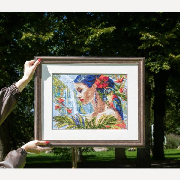A completed cross-stitch of a woman with red hibiscus flowers in her hair, a colorful parrot on her shoulder, tropical leaves in the foreground, and waterfalls in the background.
