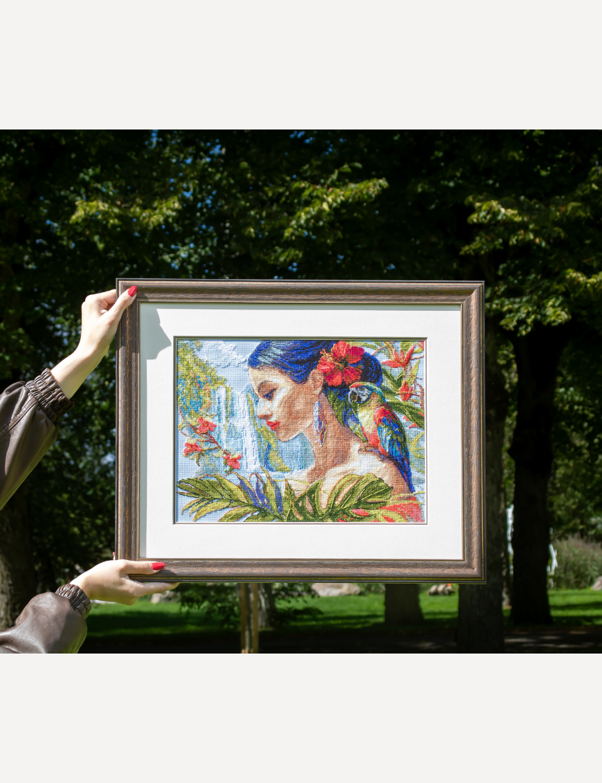 A completed cross-stitch of a woman with red hibiscus flowers in her hair, a colorful parrot on her shoulder, tropical leaves in the foreground, and waterfalls in the background.