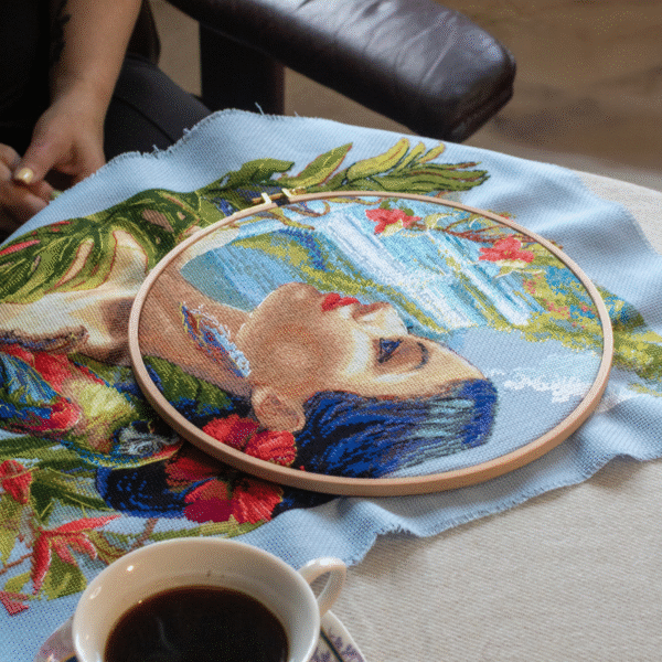 A completed cross-stitch of a woman with red hibiscus flowers in her hair, a colorful parrot on her shoulder, tropical leaves in the foreground, and waterfalls in the background.